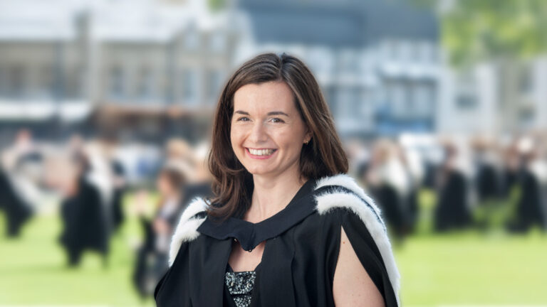 Person standing on grass in formal dress, with a group of people and buildings blurred in the background.