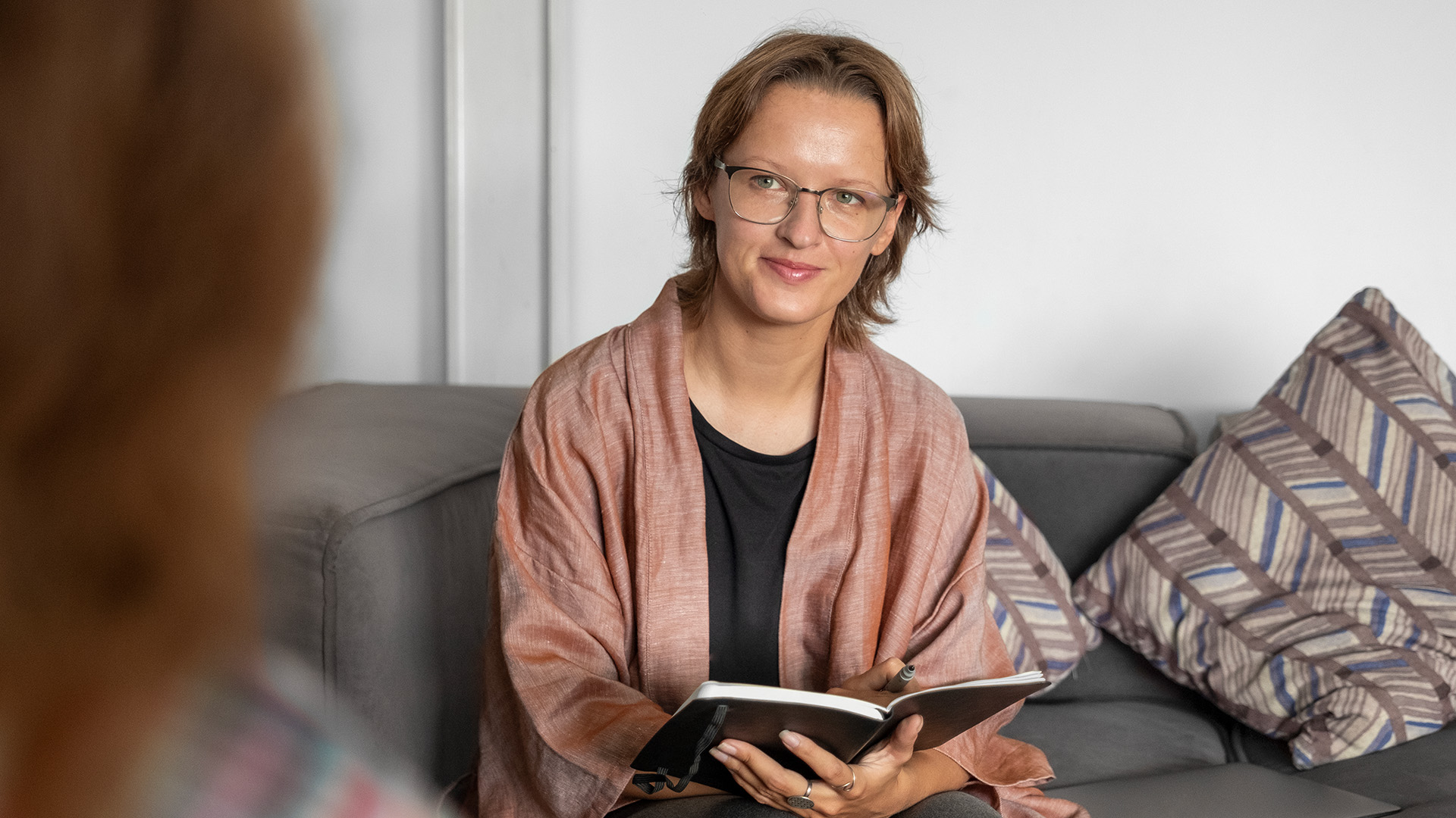 Person seated indoors with a notebook, facing another person out of focus.