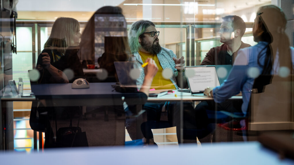 Glass‑walled meeting room showing several people seated at a table with laptops and papers.