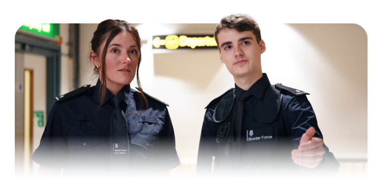 Two uniformed Border Force Officers standing in a corridor, with 'Departures' signage visible in the background.