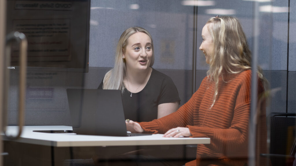 Two people seated at a table using a laptop in a meeting room.