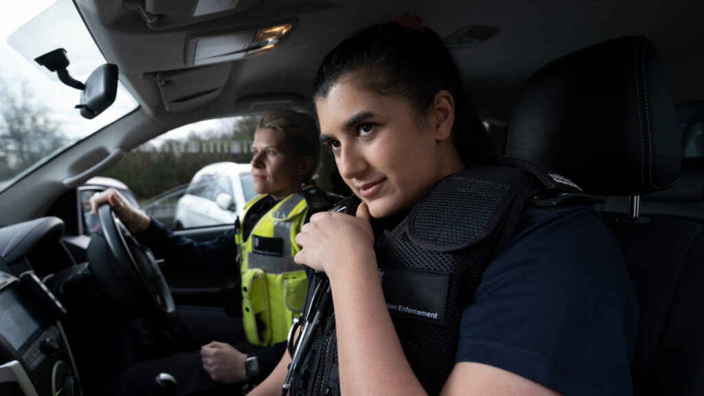Two people in uniform seated inside a vehicle, with one holding a handheld radio.