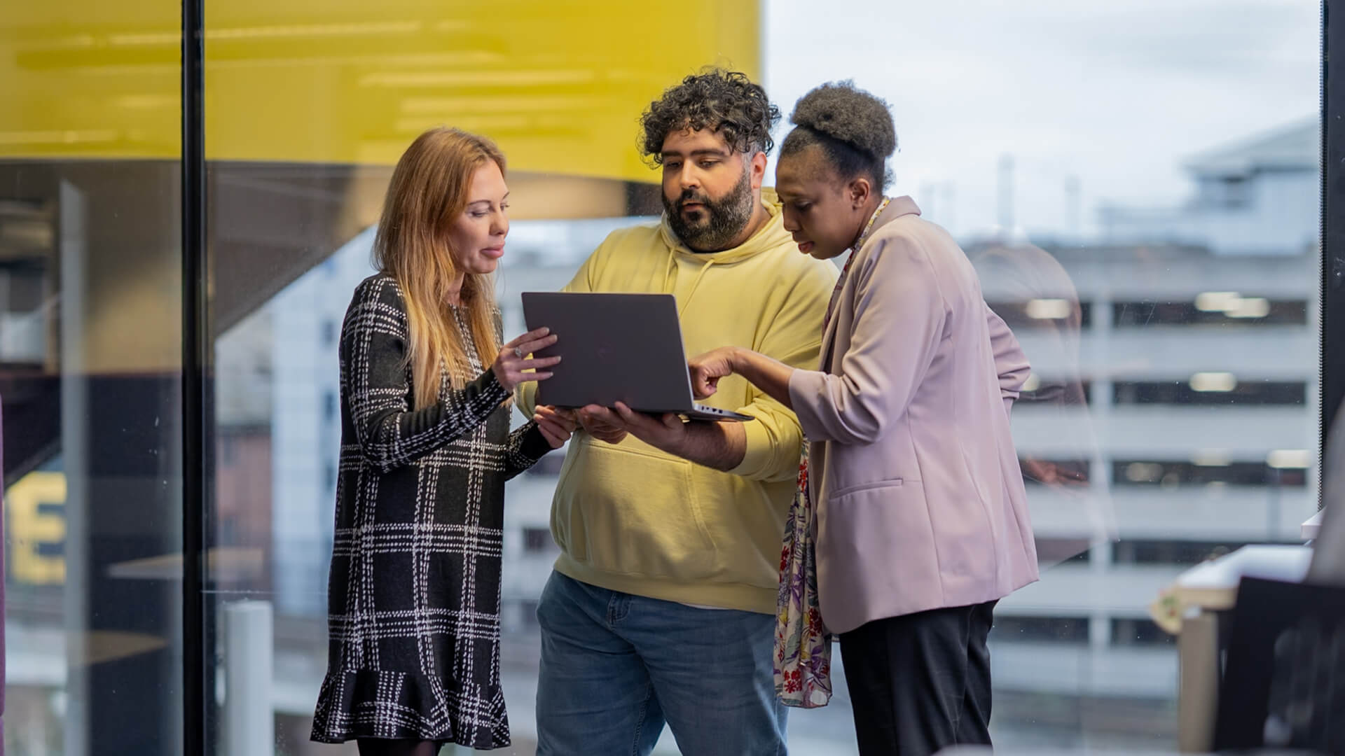 Office setting showing three people standing by a window, holding and pointing at a laptop.