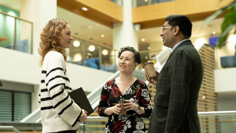 Indoor atrium with three people standing in conversation, holding takeaway cups, with stairs, seating, and upper‑level balconies visible in the background.