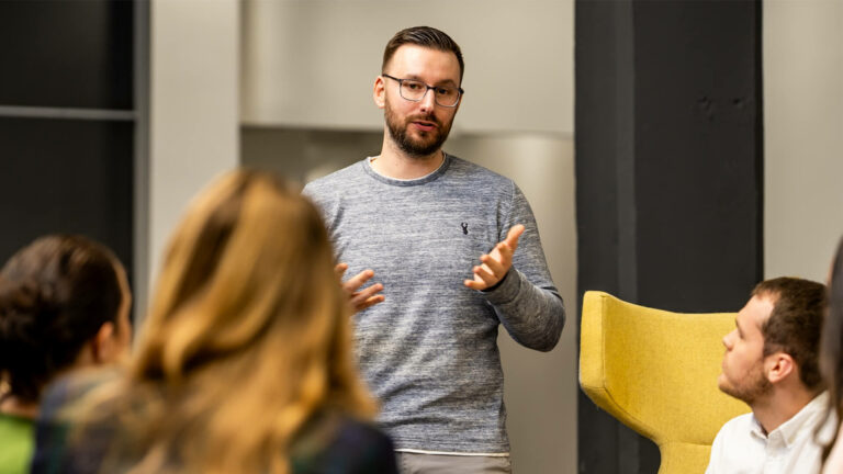 Group discussion in an indoor workspace, with one person standing and addressing others seated in chairs.