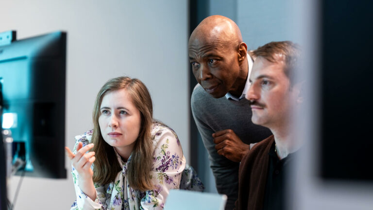 Three people collaborating at a workstation, focused on a screen.