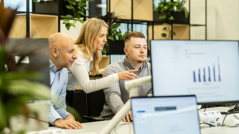 Three people gathered around a workstation reviewing information on a monitor.