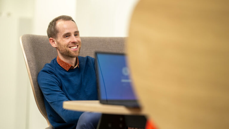 Office lounge‑style setting showing a person seated comfortably with an open laptop, viewed from the side.