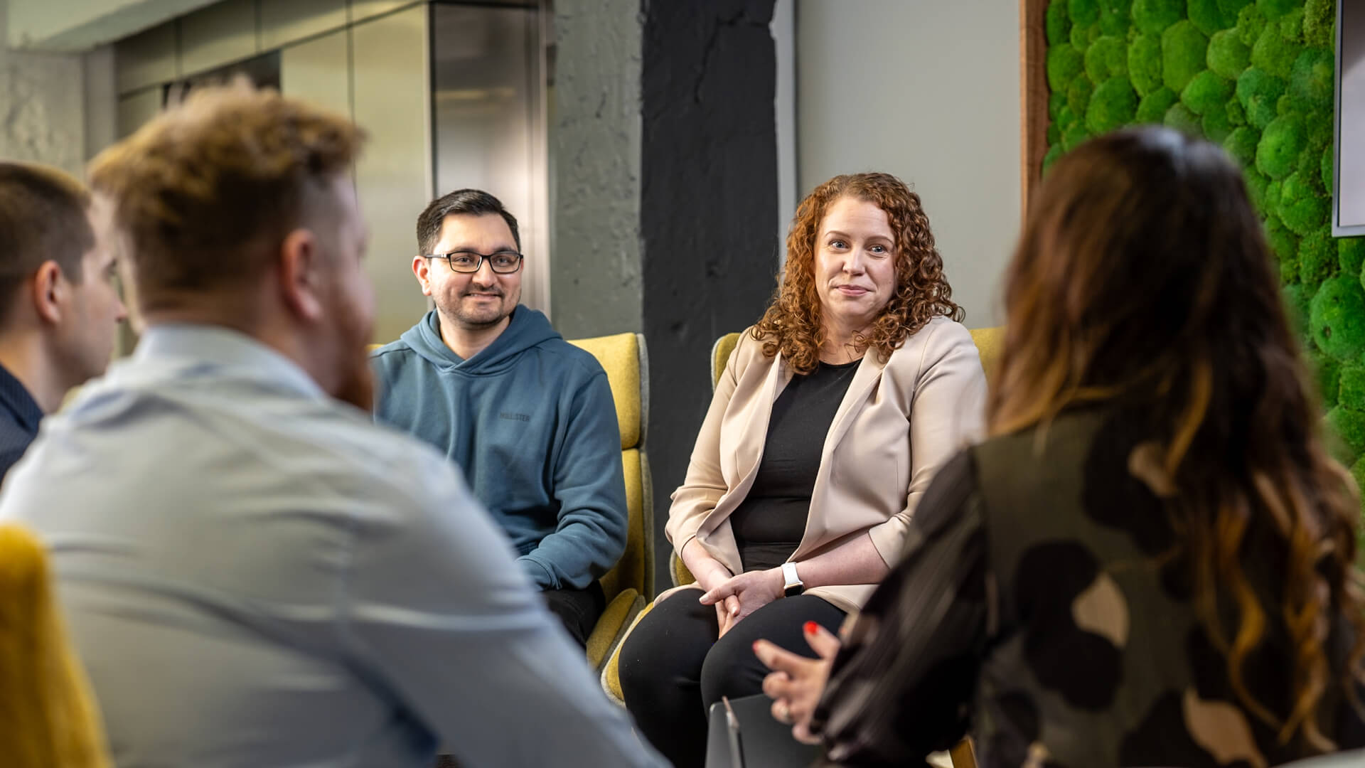 Office lounge area showing people seated and facing each other in discussion.