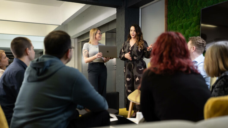 Group of people seated indoors, listening to two people standing and presenting.