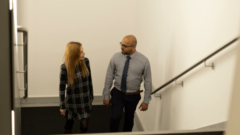 People ascending an indoor stairwell with a handrail along the wall whilst conversing.