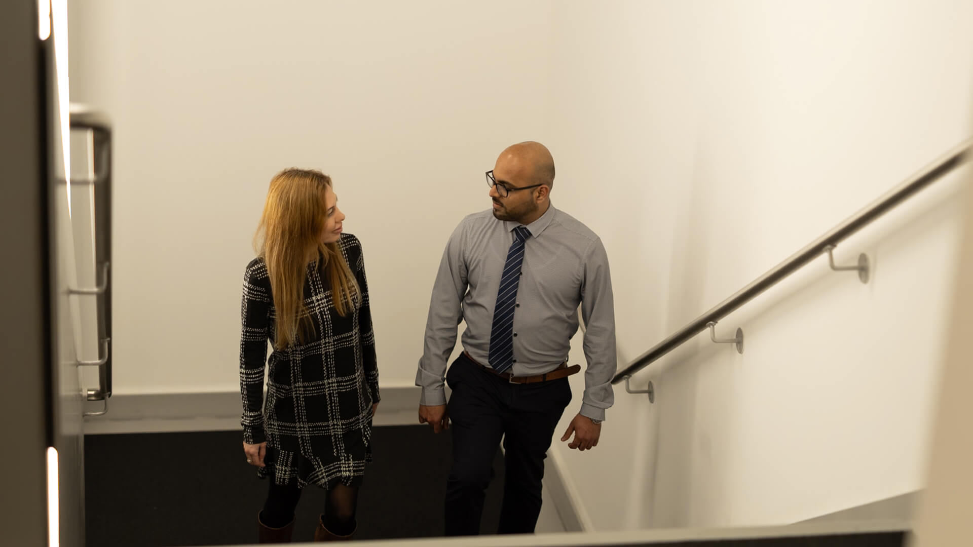People ascending an indoor stairwell with a handrail along the wall whilst conversing.