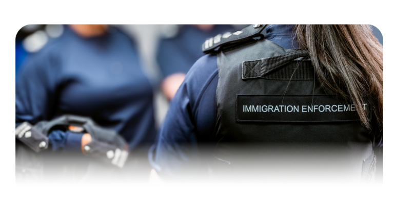 Close‑up view of a person wearing a black protective vest labelled ‘Immigration Enforcement’, with additional uniformed personnel visible nearby.