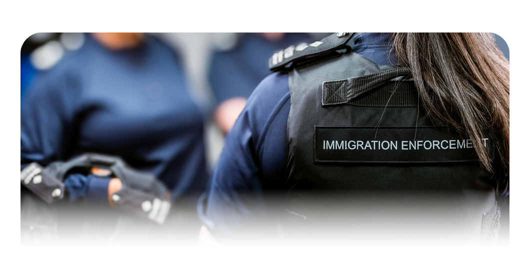 Close‑up view of a person wearing a black protective vest labelled ‘Immigration Enforcement’, with additional uniformed personnel visible nearby.